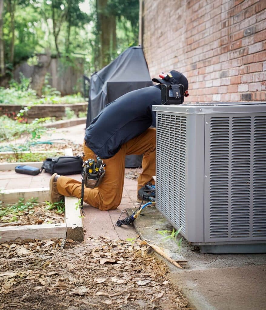 HVAC technician kneeling while repairing an air conditioning unit in a residential setting.