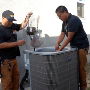 Best AC Repair: Two professional HVAC technicians are focused on repairing a Carrier brand outdoor air conditioning unit. One technician holds the fan assembly, while the other works inside the unit, emphasizing the complexity of HVAC repair. This image warns against AC repair mistakes often made by untrained individuals and promotes the use of expert teams for intricate component replacement.