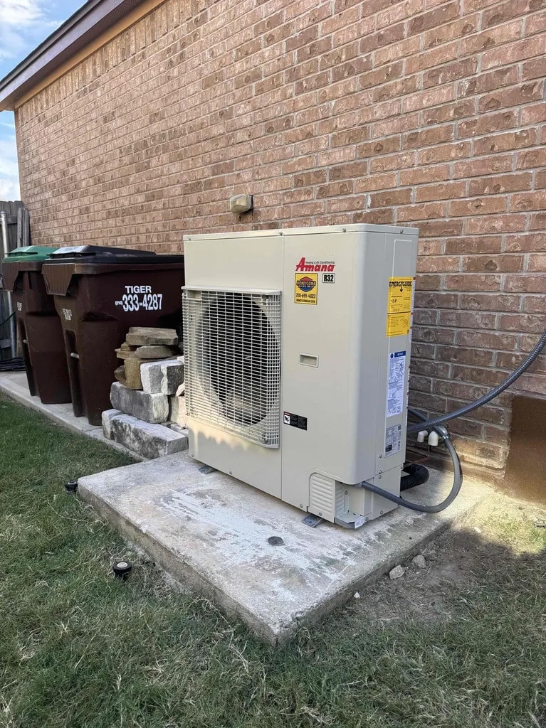 Exterior Amana inverter AC unit installed on a concrete slab alongside a brick home, representing high-efficiency system investments for inverter AC cost San Antonio, by Honeycomb.